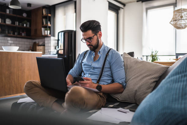 istock Modern bearded young businessman working on laptop computer while sitting on sofa at home, looking at some documents 1141259619
