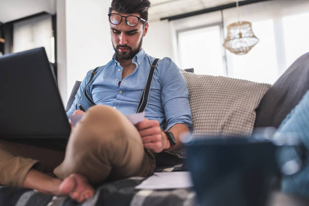 istock Modern bearded young businessman working on laptop computer while sitting on sofa at home, looking at some documents 1141259642