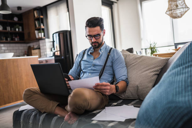 Modern bearded young businessman working on laptop computer while sitting on sofa at home, looking at some documents