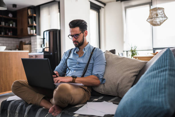 istock Modern bearded young businessman working on laptop computer while sitting on sofa at home, looking at some documents 1141260426