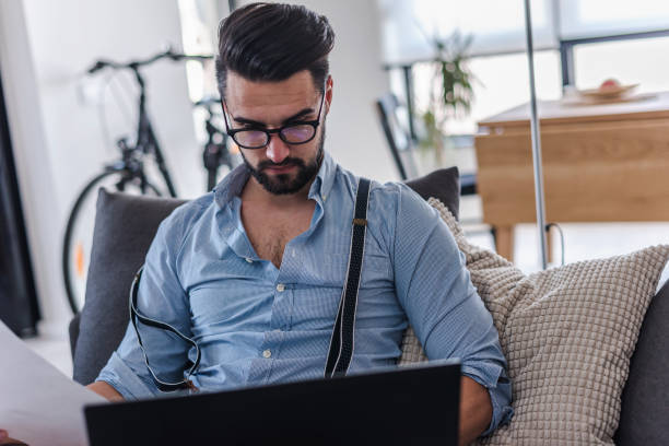 istock Modern bearded young businessman working on laptop computer while sitting on sofa at home, looking at some documents 1141260597