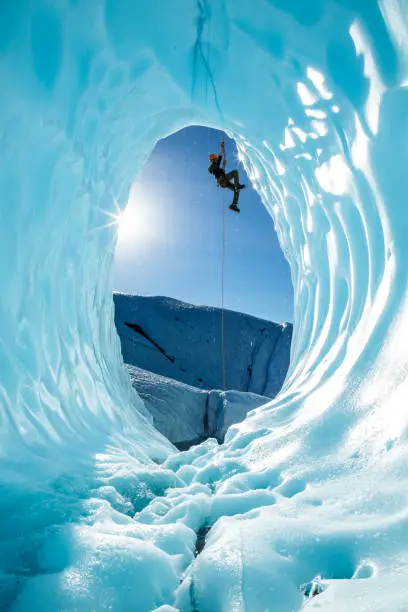 Hanging from a rope fixed above the entrance of a large blue ice cave, an ice climber ascends out of the glacial cavern. The scene is from the Matanuska Glacier, in the wilderness of Alaska.