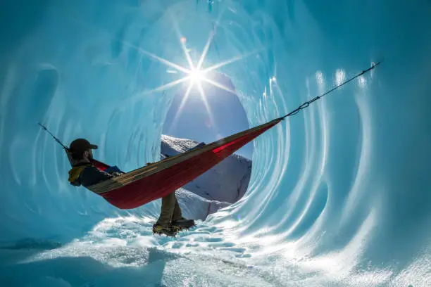 Deep in the Alaskan wilderness, an ice climber rests in a hammock set up in the walls of an ice cave. Sun shines into the entrance of the large blue cavern.