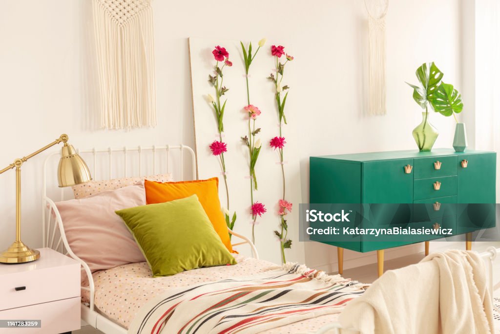 Flower board between single bed with olive green, orange and pastel pink pillows and green wooden cabinet with leaf in glass vase Bedroom Stock Photo