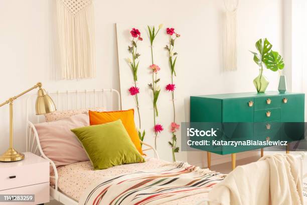 Flower Board Between Single Bed With Olive Green Orange And Pastel Pink Pillows And Green Wooden Cabinet With Leaf In Glass Vase Stock Photo - Download Image Now