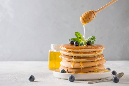 Sweet homemade stack of pancakes with pouring honey, fresh blueberry and mint in modern plate on white background. Space for text.