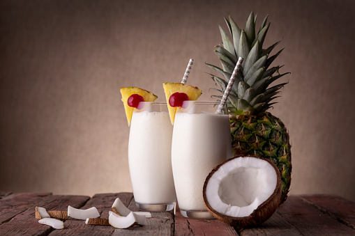 Two glasses of pina colada cocktail with dark rum, pineapple juice and coconut cream, decorated with pineapple slices and maraschino cherry on rustic wooden table