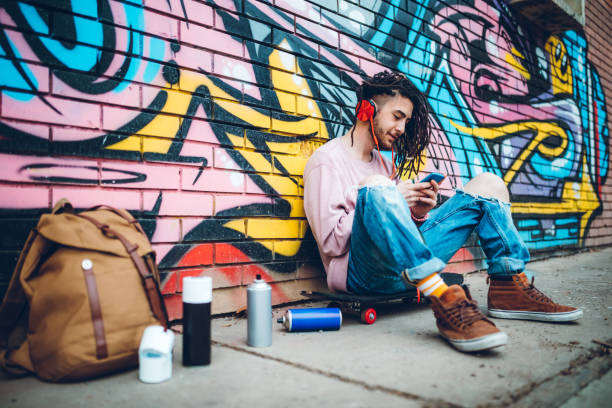 Young graffiti artist skateboarder with dreadlocks is listening to music and using phone downtown
