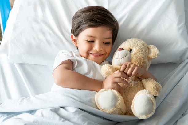 istock Playful cute boy lying down on hospital bed hugging his teddy bear 1141330665