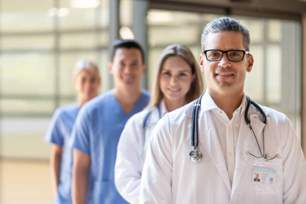 guapo practicante latinoamericano sonriendo a la cámara y equipo de pie detrás de él todos sonriendo - nurse doctor healthcare and medicine latin american and hispanic ethnicity fotografías e imágenes de stock