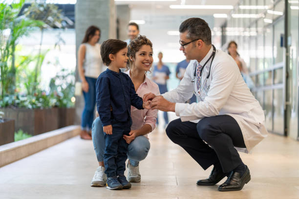 pédiatre mâle parlant à son petit patient qui est debout à côté de sa mère tout souriant - child hospital doctor patient photos et images de collection