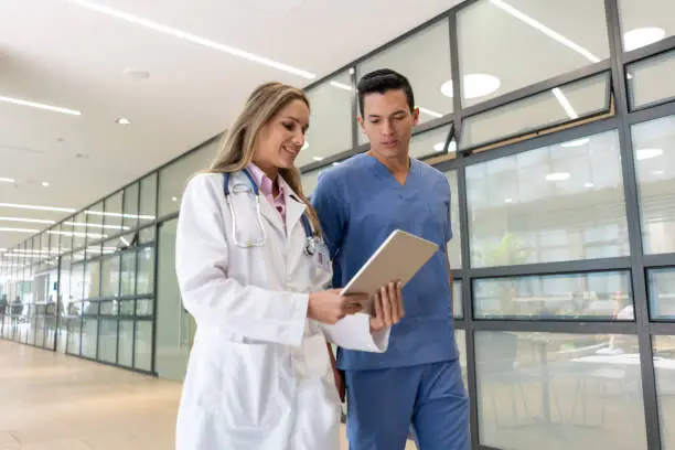 Handsome male nurse talking with female doctor while both are looking at a tablet walking through the corridor - Healthcare concepts