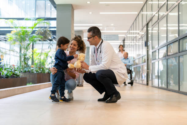 pediatra masculino jugando con un pequeño paciente usando un osito de peluche mientras mamá sonríe con mucho cariño - doctor cheerful child healthcare and medicine fotografías e imágenes de stock