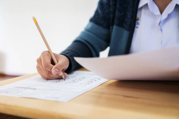 istock Students hands taking exams, writing examination room with holding pencil on optical form of standardized test with answers and english paper sheet on row desk chair doing final exam in classroom. 1141338097