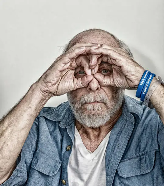 A real person, real life, senior adult man cancer chemotherapy patient is making peek-a-boo binoculars around his eyes with his cupped fingers and thumbs. He's looking forward to the day his chemotherapy ends - hoping his cancer will then be gone. The cancer themed wristbands he is wearing (personally designed/"phrased"/commissioned by me - the photographer/patient) are embossed with the words "Kick Cancer", and "Expect Success". Another day in the life of this cancer patient. Part of a "Daily Living with Cancer" image series - submitted toward brief #775310942