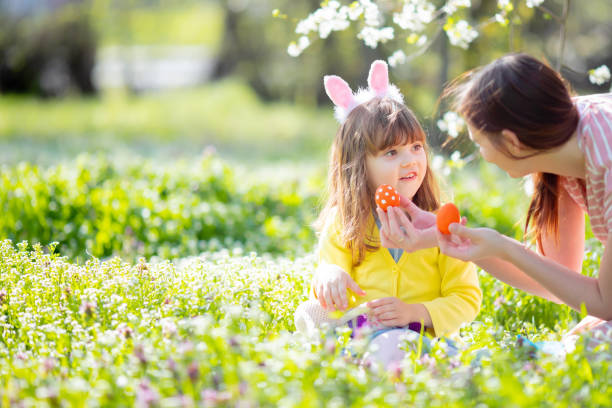バニーの耳と夏のドレスを着た巻き毛のかわいい女の子は、イースターエッグハント中に庭でリラックスし、彼女の若い母親と楽しんでいます。 - easter easter bunny fun humor ストックフォトと画像
