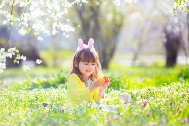 ウサギの耳と夏のドレスを着ている巻き毛のかわいい女の子は、イースターエッグハントの間に楽しんで庭でリラックス - easter easter bunny fun humor ストックフォトと画像