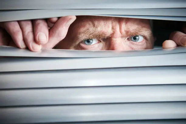 A mature man frowns, peering through closed venetian blinds nervously.