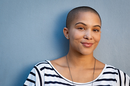 Smiling woman with bald hair looking at camera. African american woman in white and black stripe shirt standing isolated against blue background. Closeup face of proud and satisfied cool girl.