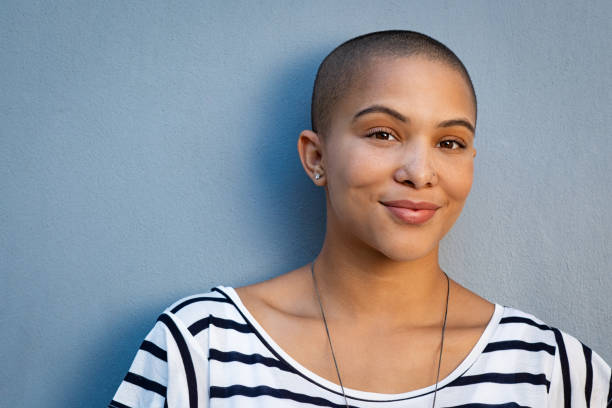 Beautiful bald woman smiling Smiling woman with bald hair looking at camera. African american woman in white and black stripe shirt standing isolated against blue background. Closeup face of proud and satisfied cool girl. balding photos stock pictures, royalty-free photos & images
