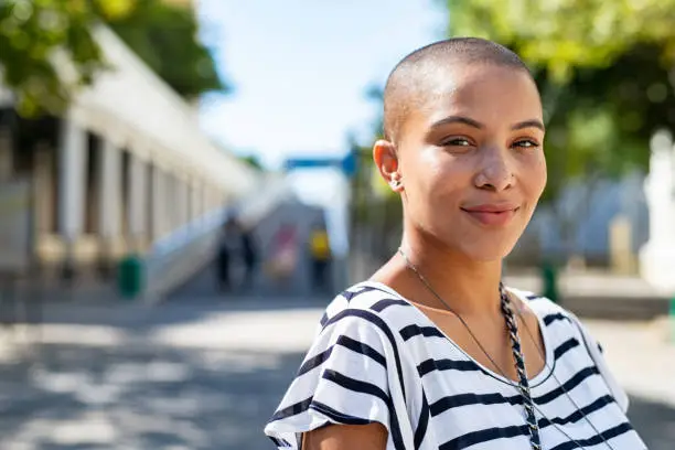 Portrait of young happy bald woman on city street looking at camera. Confident stylish girl outdoor with copy space. Proud and satisfied black curvy woman standing on street.