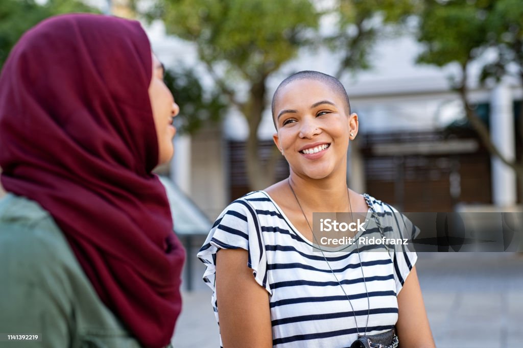 Muslim woman with friend Cheerful african american woman in conversation with muslim girl. Happy multiethnic friends in street looking at each other while in a conversation. Beautiful woman with friend in hijab talking to each others. Discussion Stock Photo