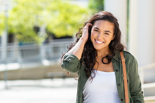 Happy young beautiful woman wearing green jacket and white shirt walking on the street. Portrait of cheerful university student looking at camera while adjusting curly hair with copy space. Latin stylish girl smiling while standing on street.