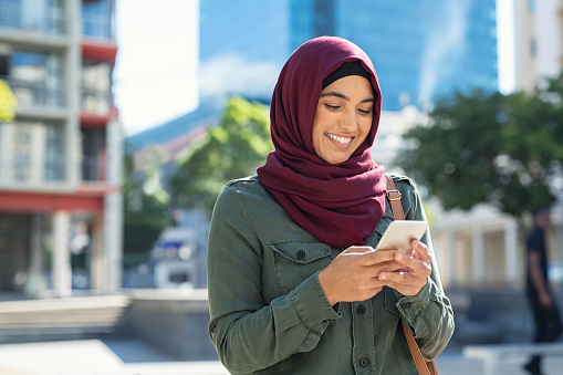 Pretty muslim woman using mobile phone outdoor. Arabic woman wearing hijab using smartphone on the street. Islamic girl texting a message phone in city centre.