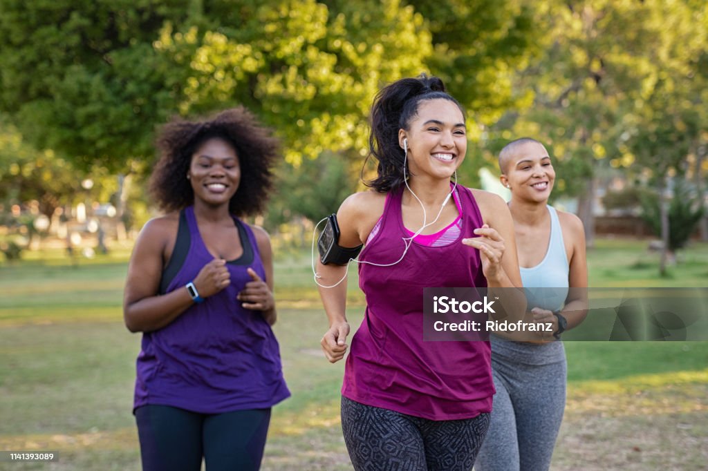 Active curvy women jogging Group of curvy girls friends jogging together at park. Beautiful smiling young women running at the park on a sunny day. Female runners listening to music while jogging. Exercising Stock Photo