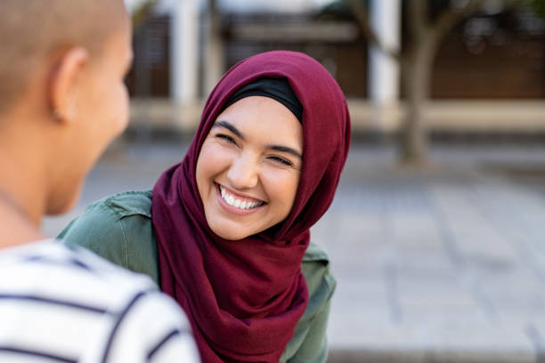 Multiethnic friends in conversation Portrait of cheerful woman in hijab smiling while talking to bald friend. Best friends on street laughing. Back view of beautiful islamic woman with toothy smile looking at friend outdoor. chubby arab stock pictures, royalty-free photos & images