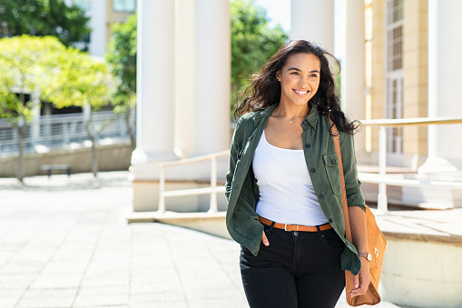 Pretty casual natural woman walking in urban street and looking away. Cheerful latin girl with bag travelling across the city. Beautiful young woman smiling outdoor.