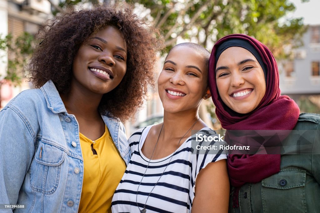 Multiethnic young friends enjoying together Group of three happy multiethnic friends looking at camera. Portrait of young women of different cultures enjoying vacation together. Smiling islamic girl with two african american friends outdoor. Multiracial Group Stock Photo