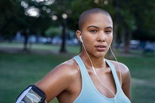 Portrait of motivated young woman resting after jogging. Beautiful focused bald girl take a break after run. Determined runner listening to music while jogging and looking at camera.