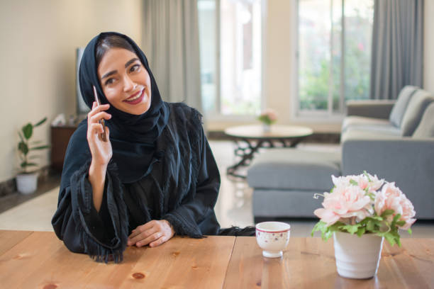 jeune femme musulmane de sourire parlant sur le téléphone mobile tout en appréciant le café du matin à la maison - desk on the phone sitting table photos et images de collection