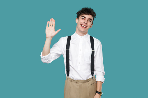 Happy excited man toothy smile and greeting. Portrait of handsome hipster curly young businessman in classic casual white shirt and suspender standing. indoor studio shot isolated on blue background.