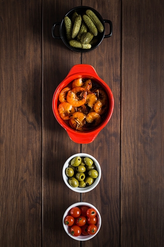 Four tapas dishes in a vertical column on a wooden table, including gherkins, garlic prawns, olives and cherry tomatoes.
