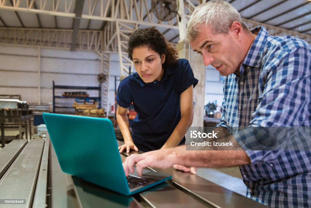 Instructor explaining female trainee over laptop Mature instructor explaining female apprentice over laptop. Trainee is listening to engineer at factory. They are working in industry. Laptop Stock Photo
