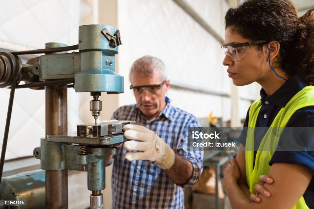 Instructor teaching female trainee in factory Mature instructor teaching female trainee to use yoke machine. Engineer is with apprentice in industry. They are in factory. Industry Stock Photo