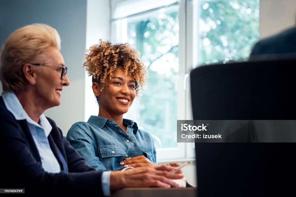 Smiling businesswomen looking away in office Smiling businesswomen looking away while sitting at desk in office. Female professionals are with confident look on their faces. They are working on start-up business. 35-39 Years Stock Photo