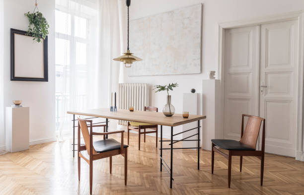 istock Stylish and modern dining room interior with design sharing table, chairs, gold pendant lamp, abstract paintings and elegant accessories. Tropical leafs in vase. Eclectic decor. Brown wooden parquet. 1141409300