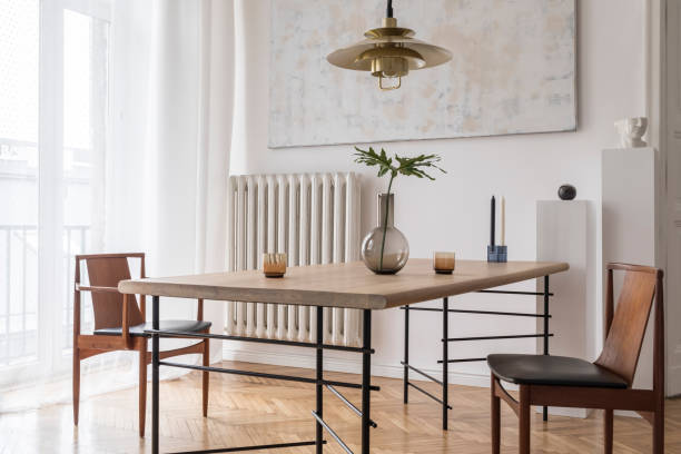 istock Stylish and modern dining room interior with design sharing table, chairs, gold pendant lamp, abstract paintings and elegant accessories. Tropical leafs in vase. Eclectic decor. Brown wooden parquet. 1141409340
