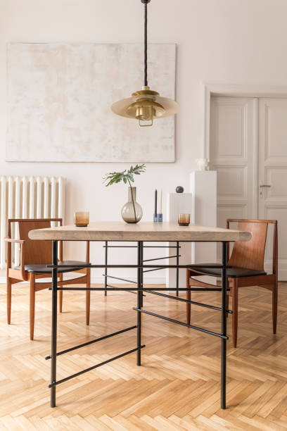 istock Eclectic and elegant dining room interior with design sharing table, chairs, gold pedant lamp, abstract paintings and stylish accessories.Tropical leafs in vase. Minimalistic decor. Real photo. 1141409341