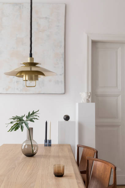 istock Eclectic and elegant dining room interior with design sharing table, chairs, gold pendant lamp, abstract paintings and stylish accessories. Tropical leafs in vase.Eclectic decor. Brown wooden parquet. 1141409344