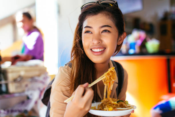 Young female tourist eating pad thai noodle at the shop Young Asian tourist woman eating pad thai noodle, traditional street food in Thailand and looking far away thai ethnicity stock pictures, royalty-free photos & images