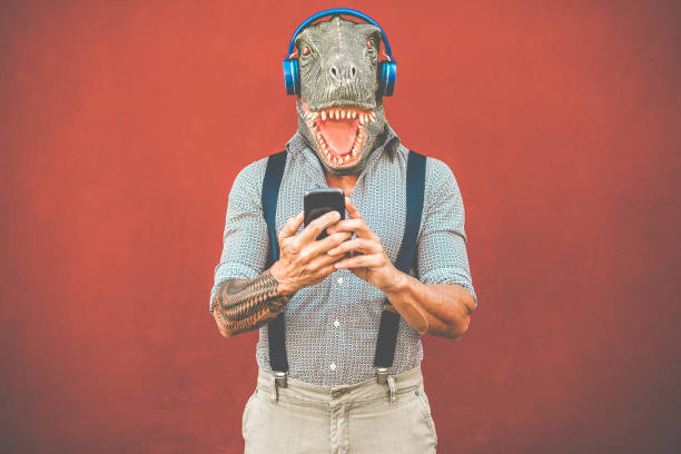 istock Tattooed man with t-rex mask using smartphone while listening music - Crazy senior guy choosing playlist from mobile phone app - Technology trends and madness costume concept - Focus on face 1141427197