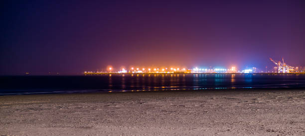 de kustlijn van blankenberge strand verlicht door de nacht, kleurrijke verlichting op de industrieterrein in de verte - blankenberge strand stockfoto's en -beelden