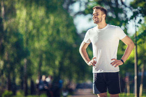 Happy morning. Cheerful young bearded man in sport clothes is standing and keeping arms on his hips while looking aside with wide smile. He is enjoying warm summer day. Copy space in the left side