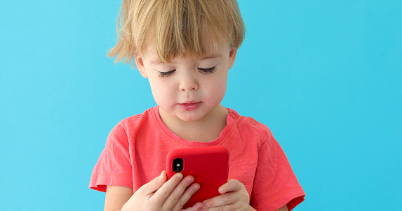 Concentrated child tapping cell phone screen, interest in modern technology. Pretty boy blue background