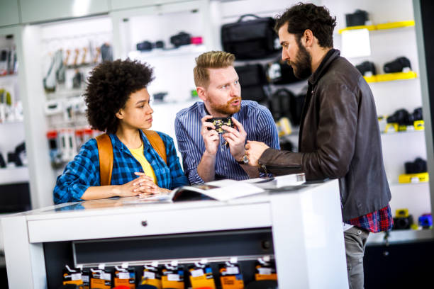 istock Store seller holding a small compact camera with a camo pattern and helping a young buyer couple by explaining it's functionalities 1141457059