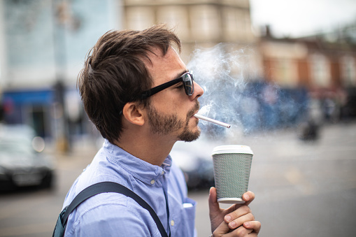 Young man with sunglasses smoking cigarette  and drinking coffee on the street in London